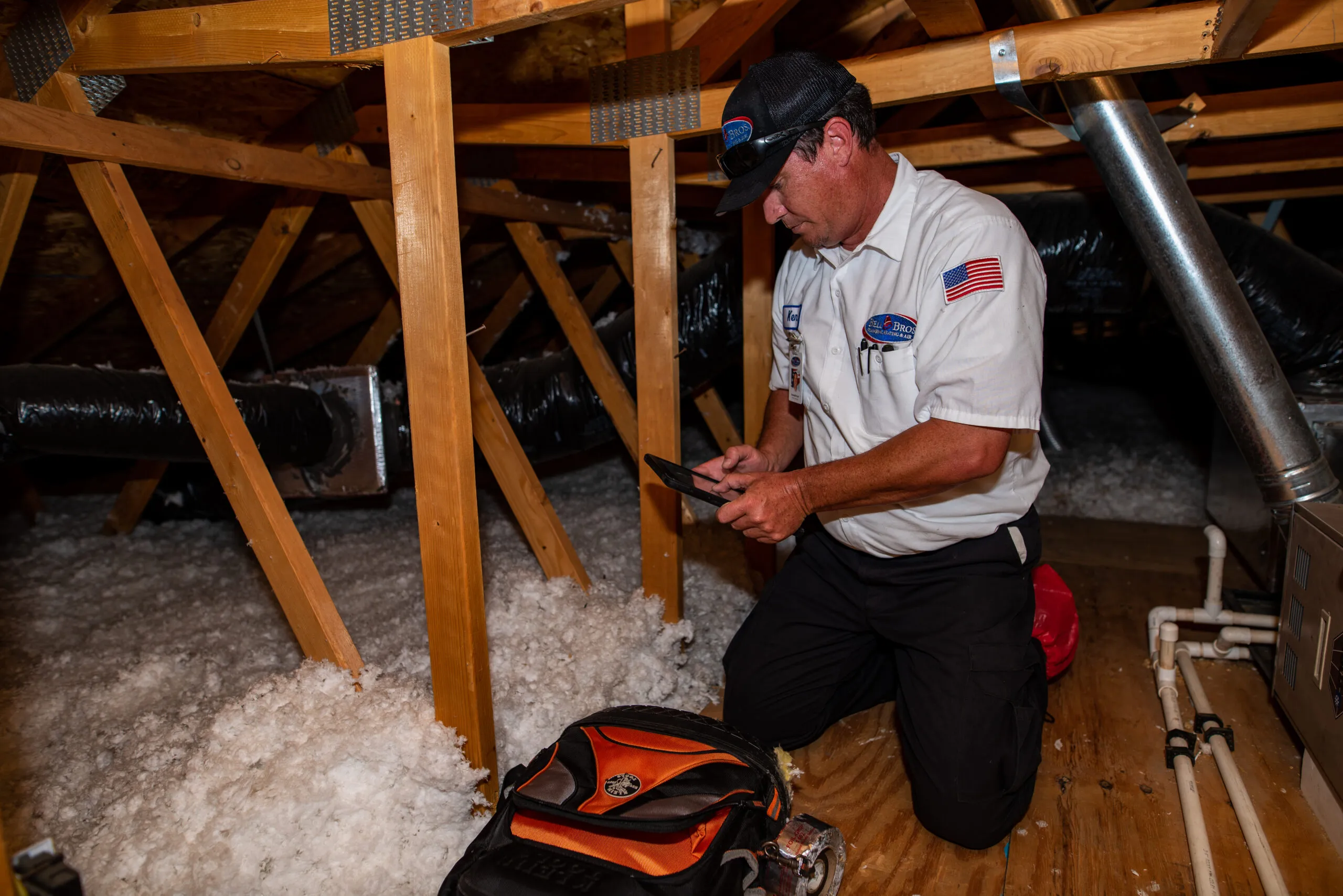 A technician from BellBrothersPlumbingHeatingandAir in Brea kneels in an attic, working on a heating system with tools and insulation visible around him.